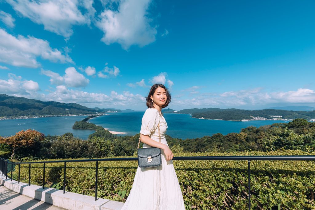 An Asian woman stands by a railing overlooking a stunning ocean view under a clear blue sky.